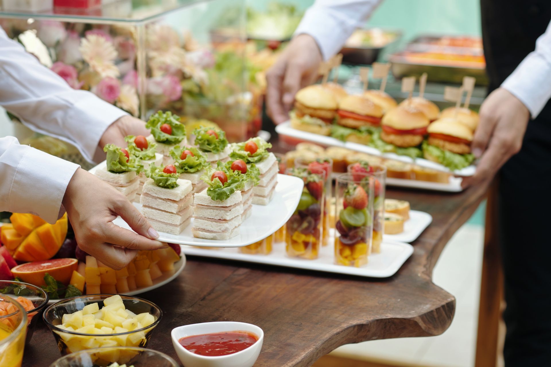 Servers arrange various gourmet finger foods and sandwiches on a rustic wooden buffet table.