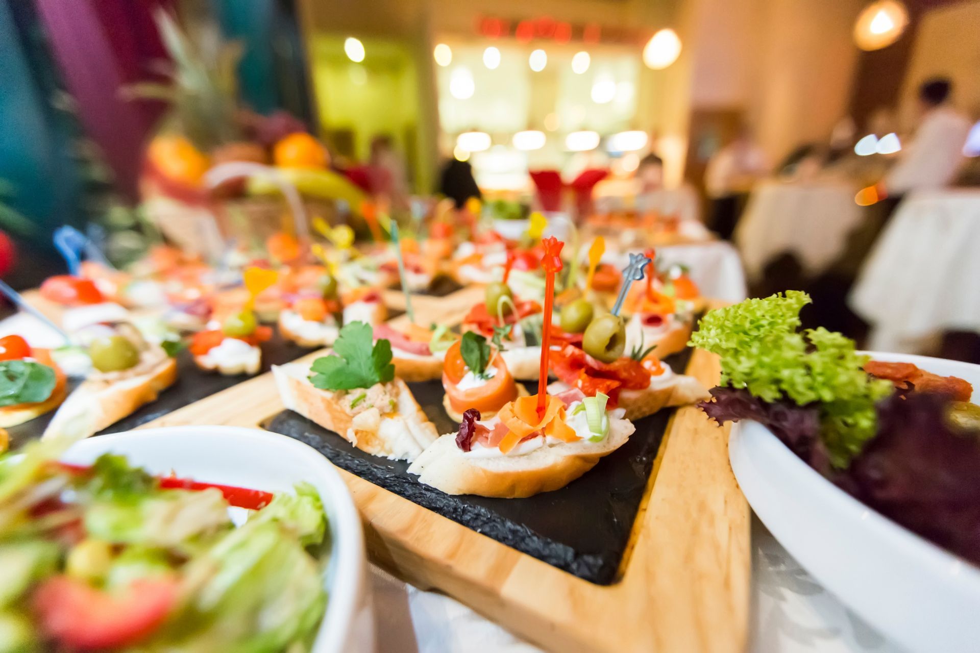 A variety of appetizers and small sandwiches displayed on stone boards at a buffet, with a blurred dining room background.