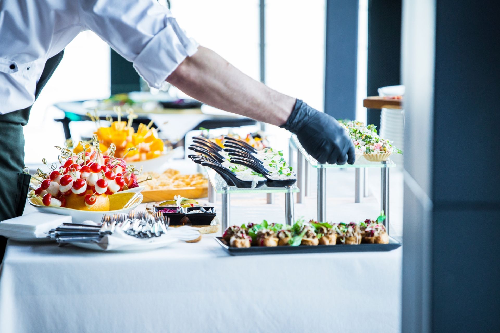 A person wearing a black glove places hors d'oeuvres on a buffet table set with various finger foods and white tablecloths.