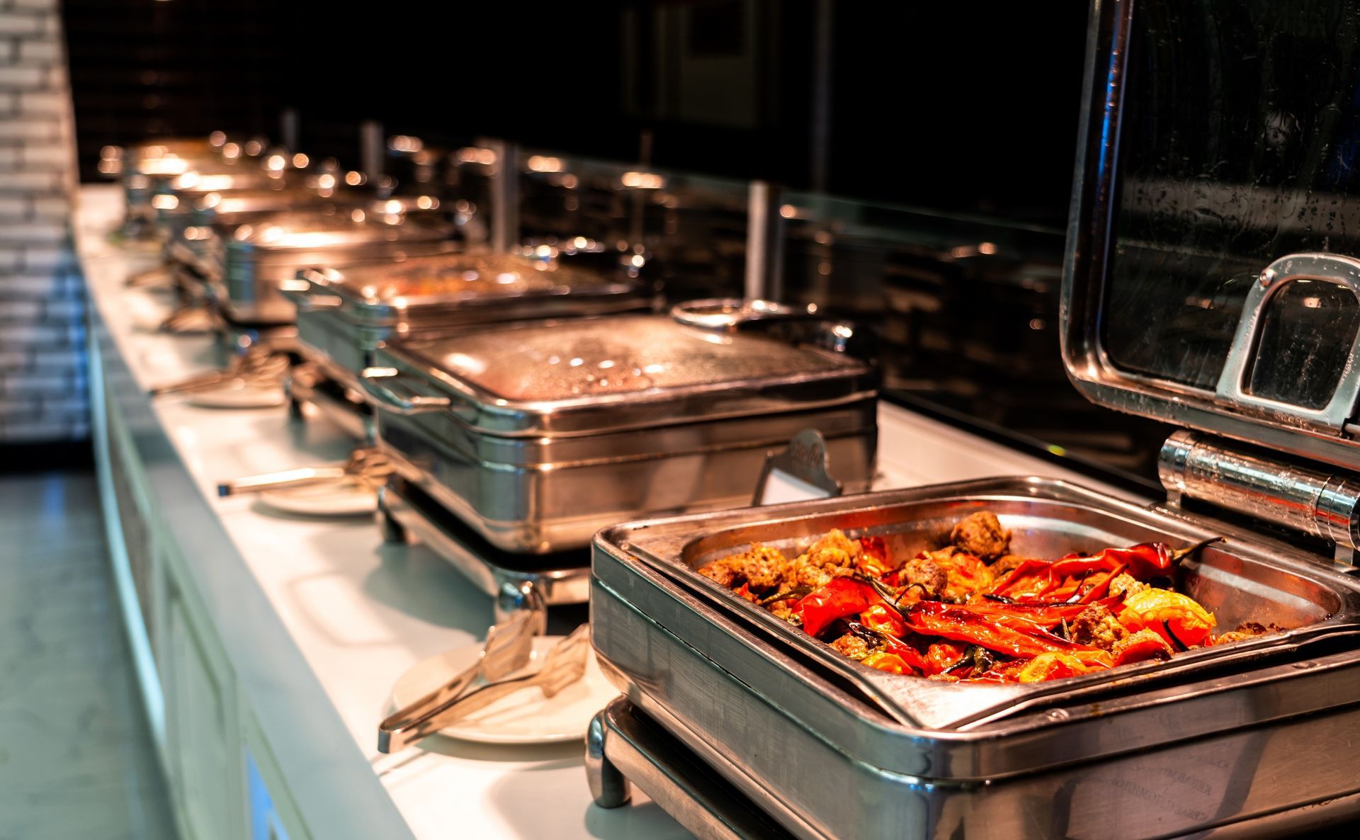 A row of stainless steel chafing dishes on a buffet line, with the closest container filled with cooked food.