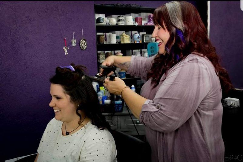 Stylist curling a woman’s hair in a salon with purple walls and shelves in the background