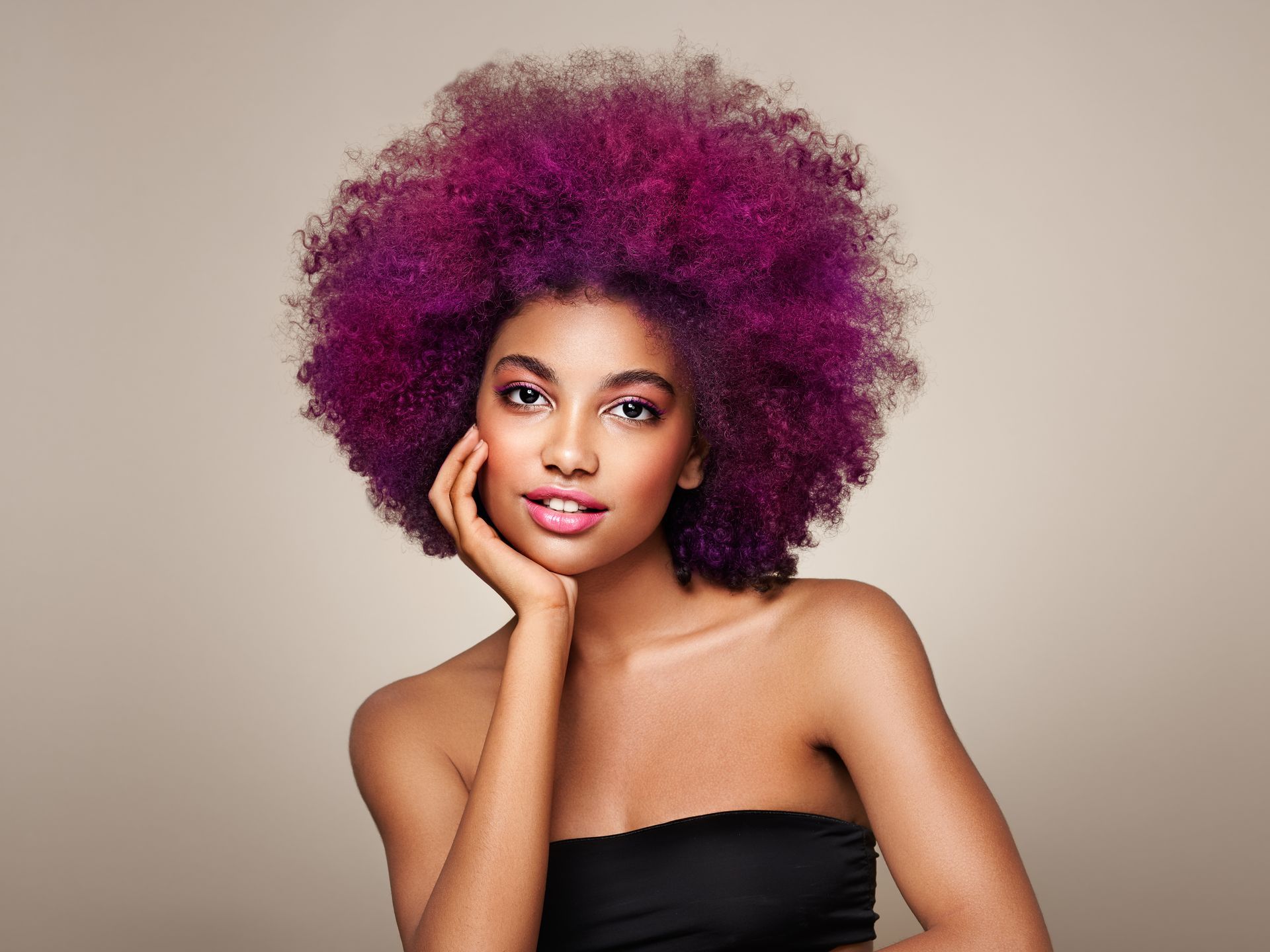 Portrait of a person with a voluminous purple afro, resting their chin on their hand against a neutral background