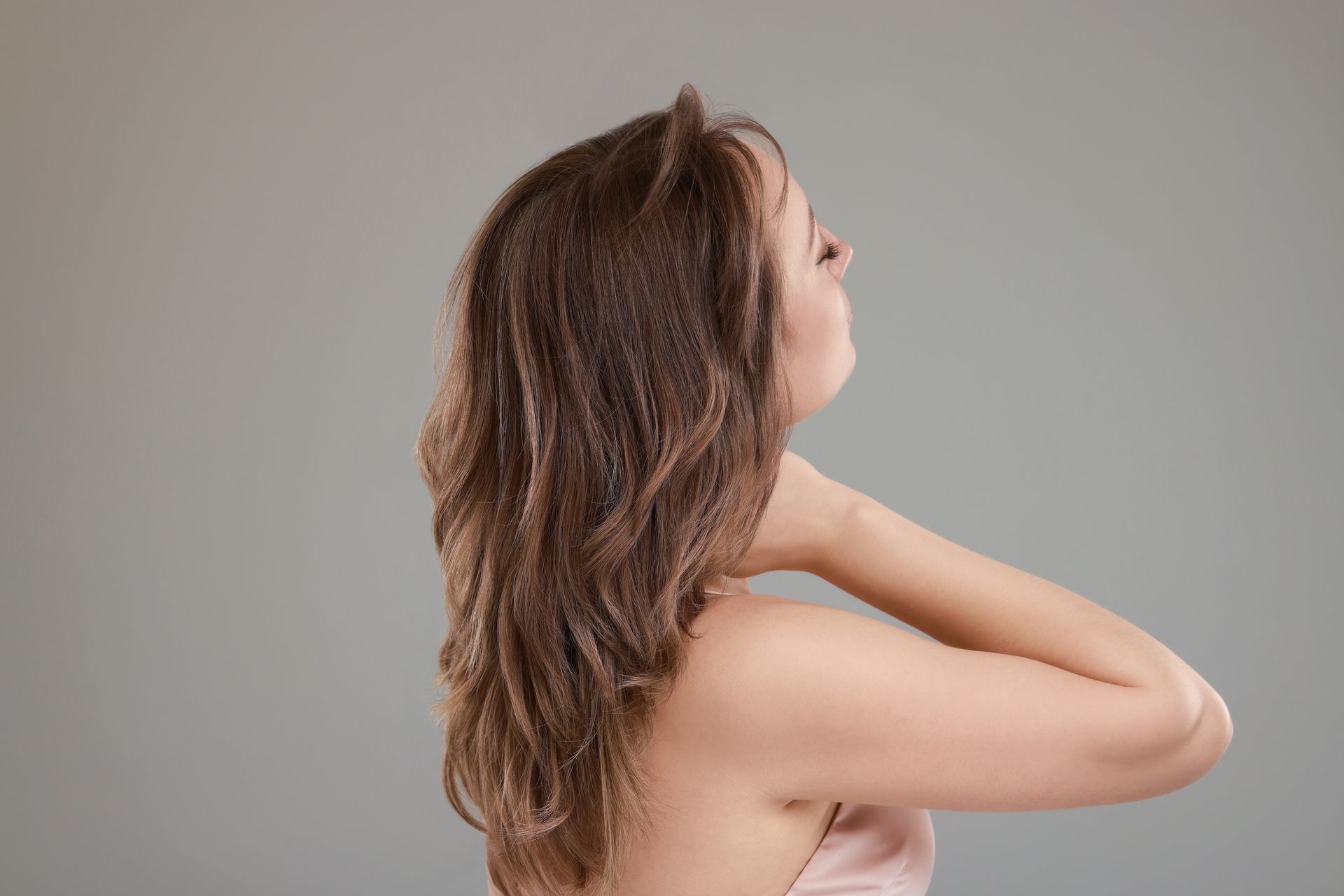 Side-profile of a person with long brown hair touching the back of their neck against a gray background