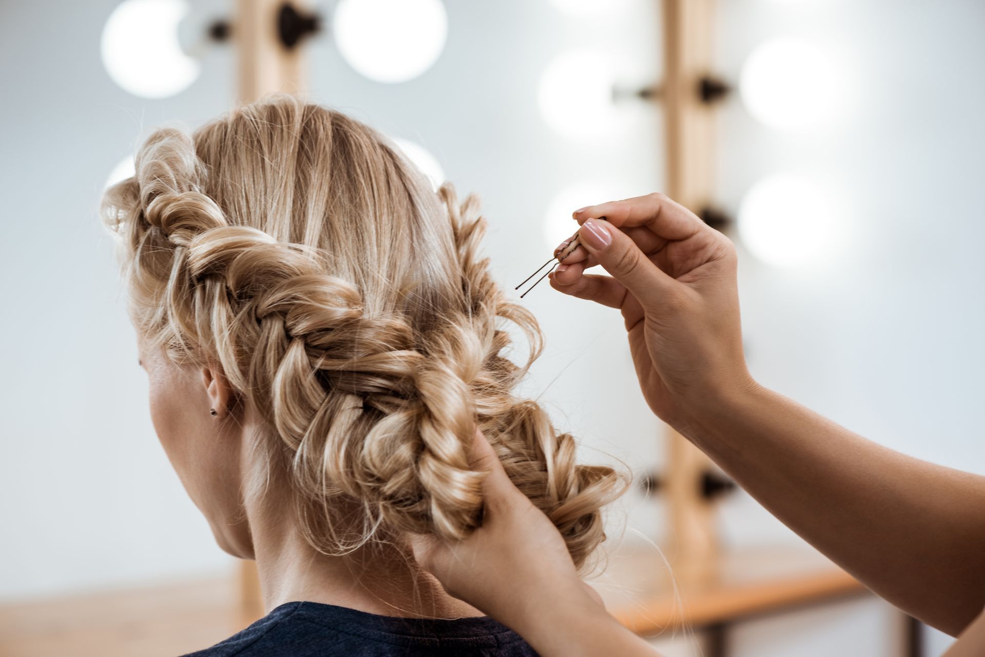 Braided blonde hair being styled by a hand in front of a mirror and vanity lights