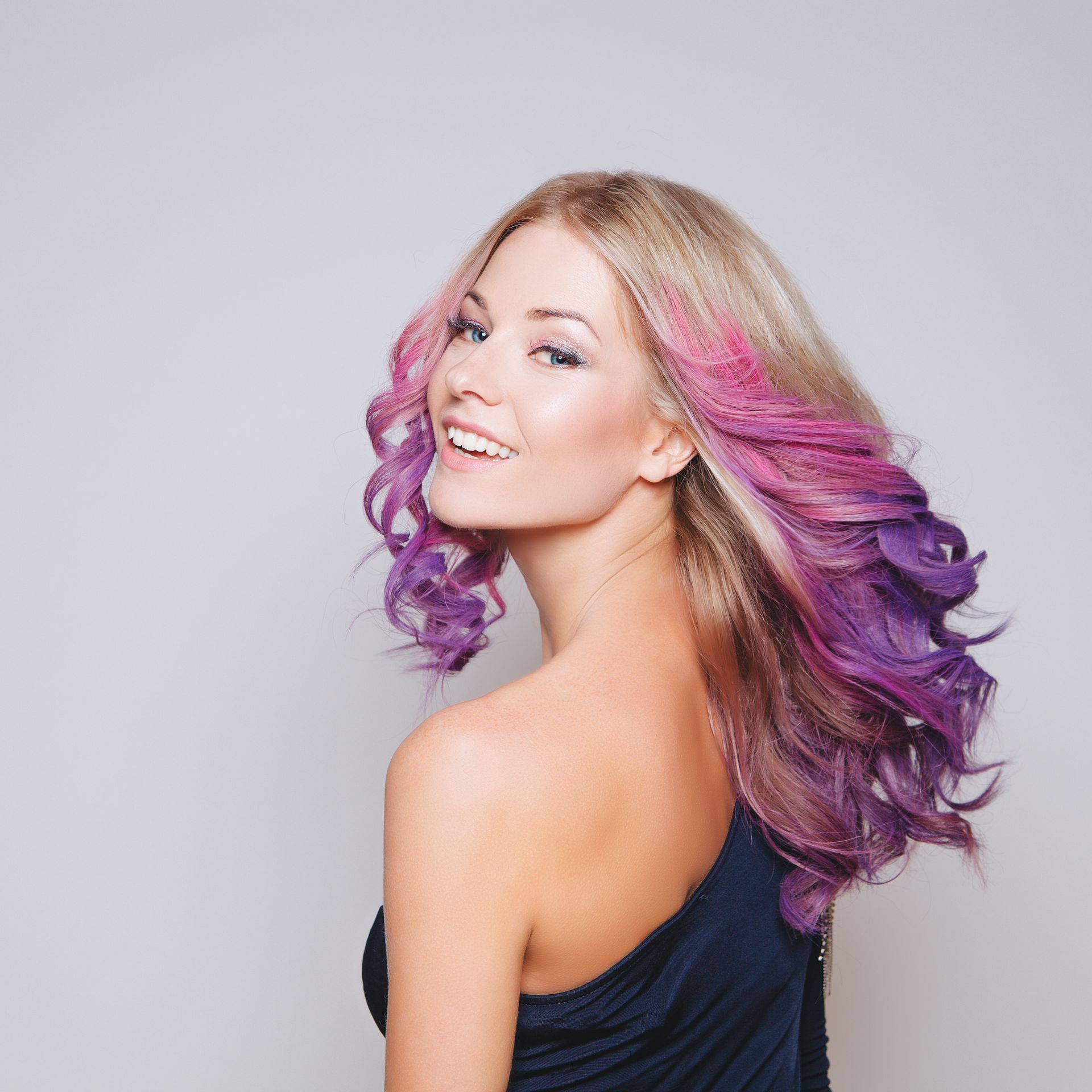 Woman with long pink and purple hair smiling over her shoulder against a gray background