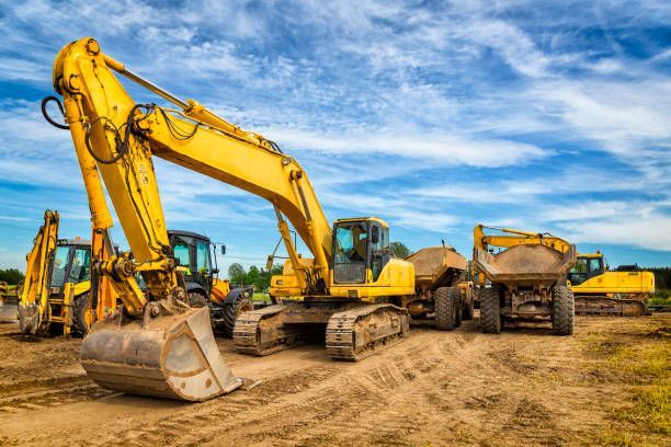 Yellow excavators and dump trucks parked on a dirt construction site under a cloudy blue sky. Yellow excavators and dump trucks parked on a dirt construction site under a cloudy blue sky.
