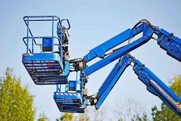 Two blue aerial work platforms, one elevated above the other, against a clear blue sky and trees. Two blue aerial work platforms, one elevated above the other, against a clear blue sky and trees.