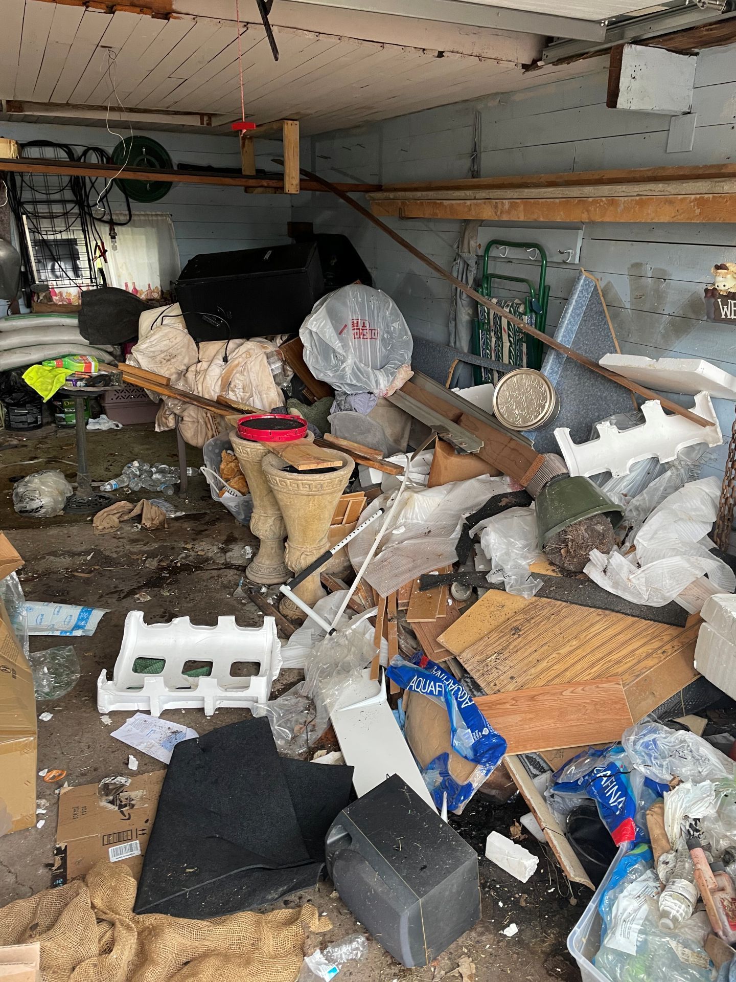 Cluttered garage packed with debris, broken furniture, boxes, and scattered household items.
