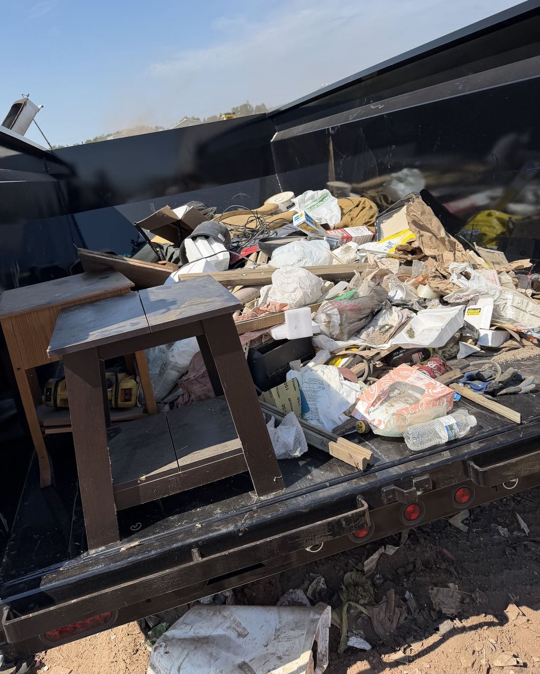 Overflowing dumpster filled with mixed trash and debris, with a small wooden stool in front