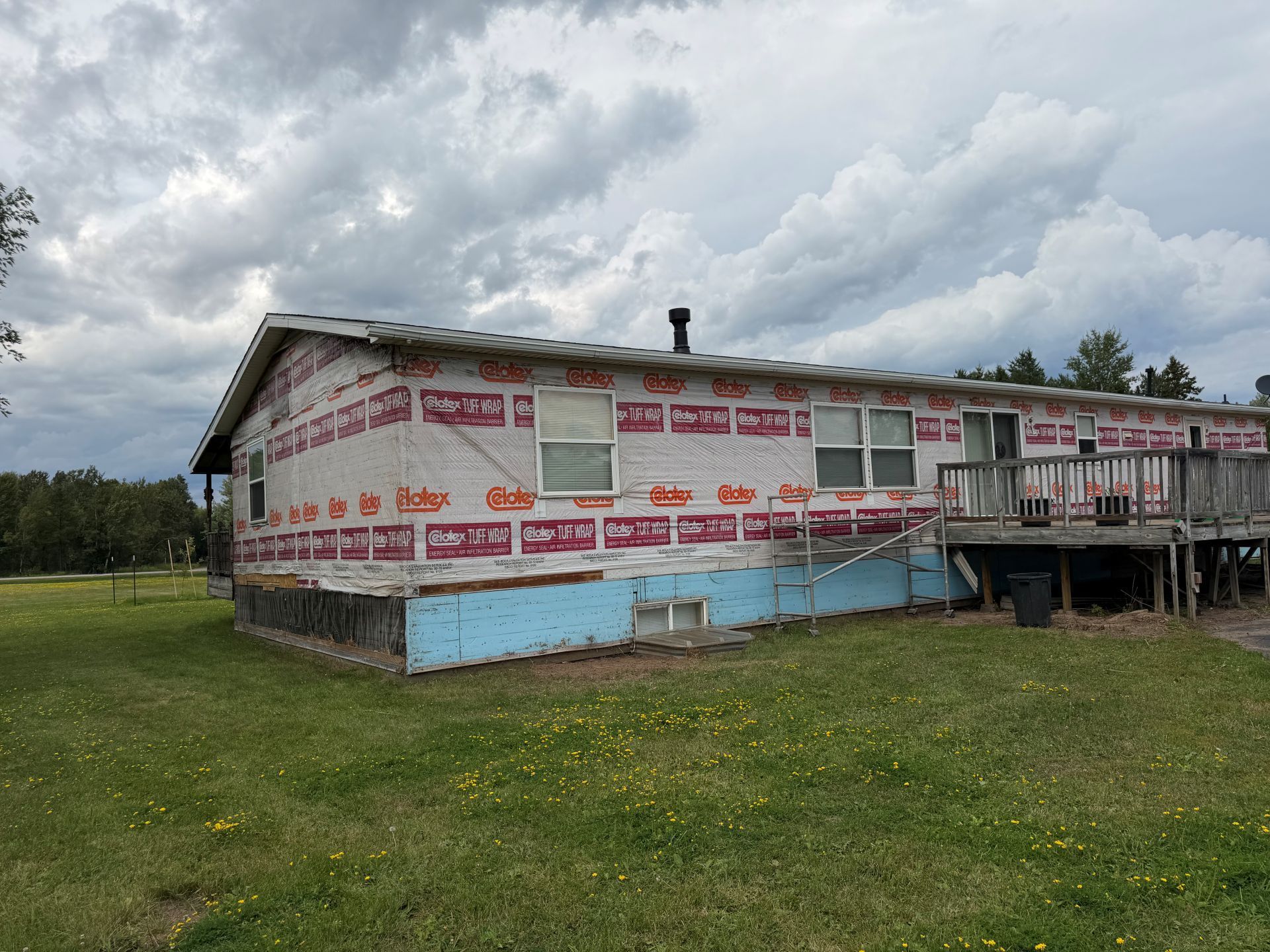 Old weathered house with red trim, wrapped in insulation, set on a grassy lot under cloudy skies