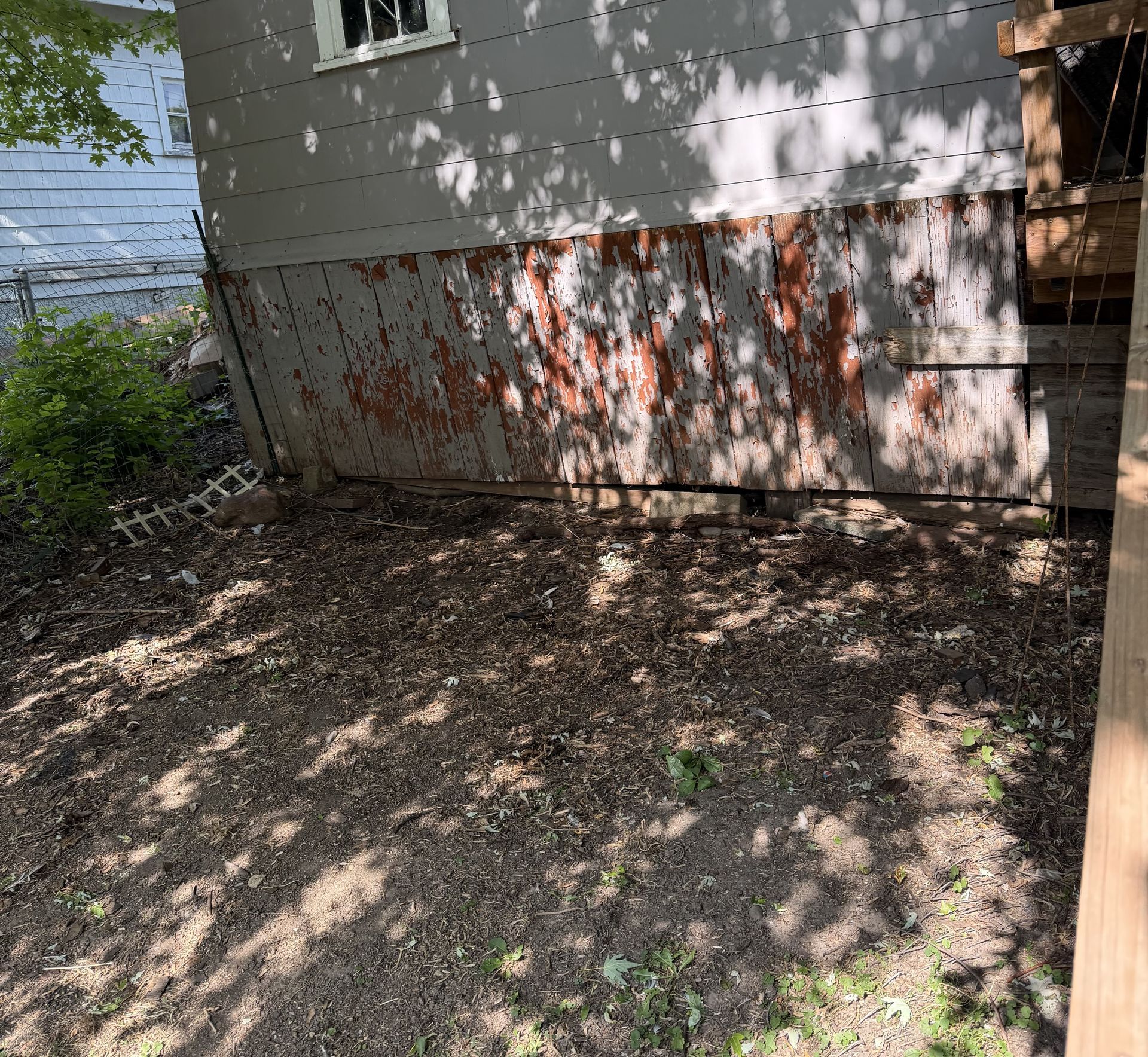 Shaded backyard beside a house with dirt ground, trees, and a wooden fence.
