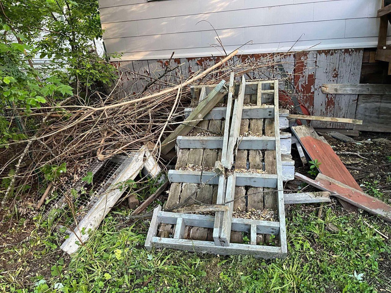 Piles of storm-damaged debris and broken wooden frames beside a house wall and overgrown yard