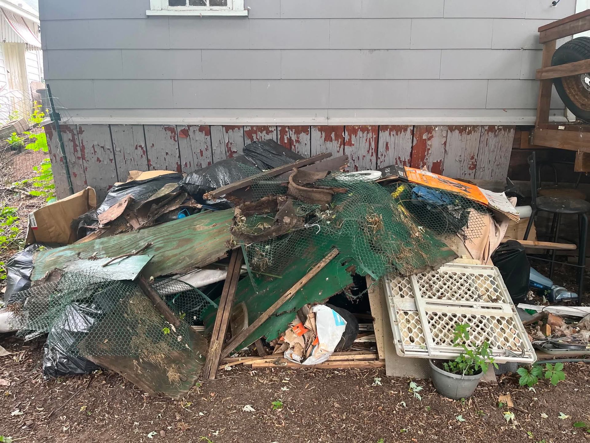 Pile of debris and scrap wood, with a green tarp and crate, stacked against a house exterior.