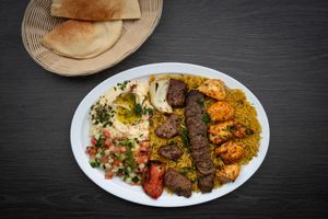 A plate of food with rice , meat , vegetables and hummus on a table.