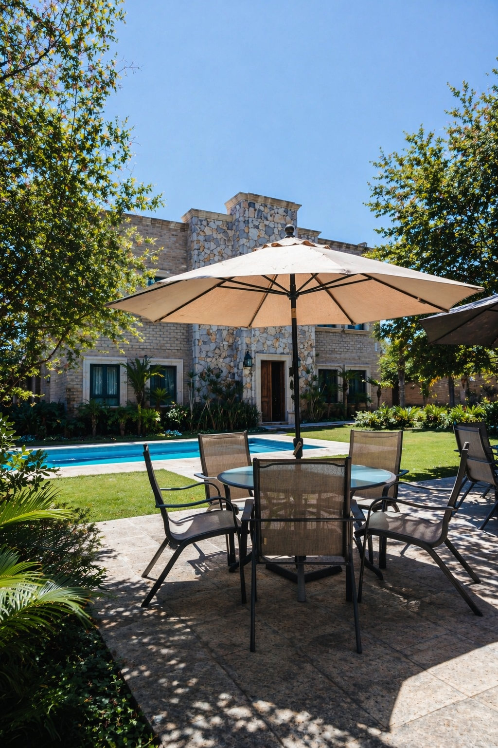 Un patio con un conjunto de comedor redondo bajo una gran sombrilla beige, con vistas a una piscina y una casa de piedra.