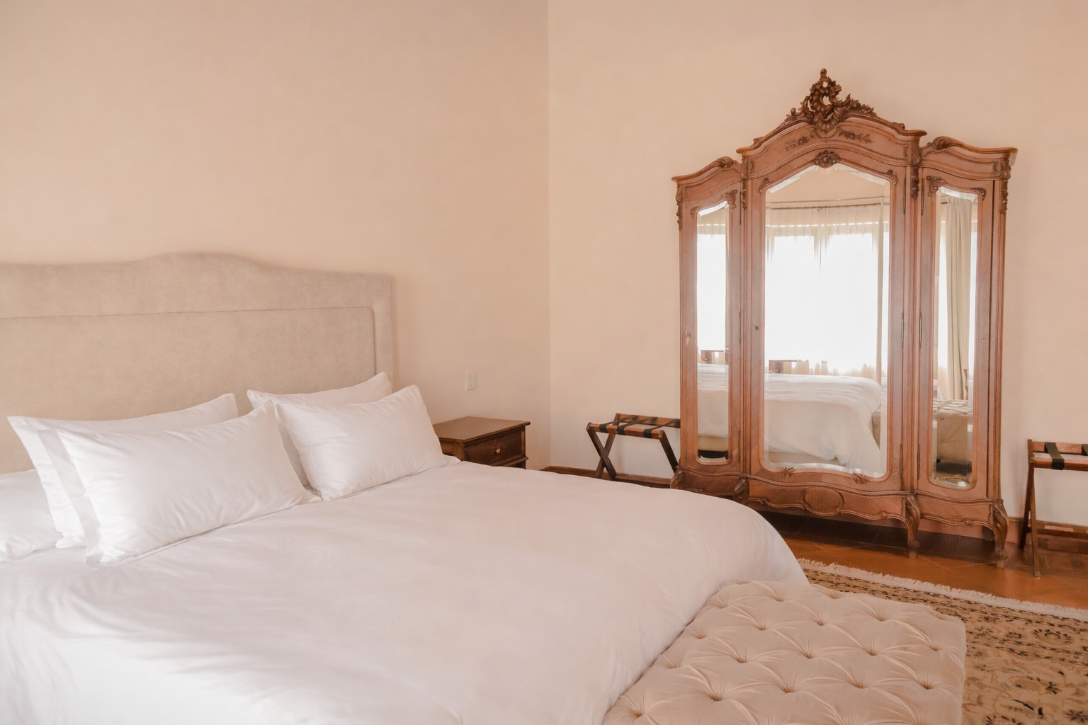 Un dormitorio con una cama que cuenta con un cabecero de color claro, ropa de cama blanca impecable y un gran espejo de tocador de madera ornamentado.