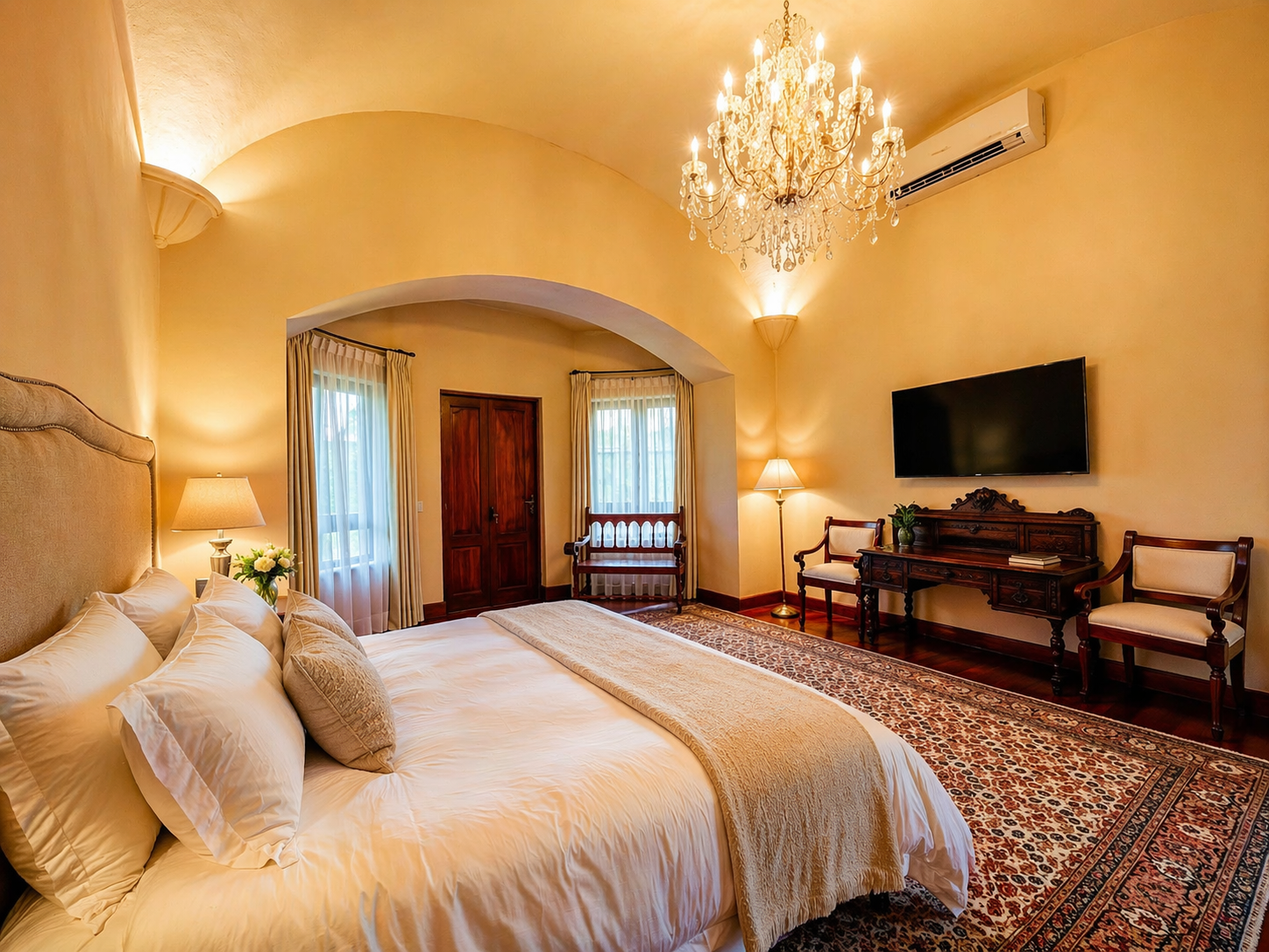 Una lujosa habitación de hotel con una cama grande, una lámpara de araña de cristal, paredes arqueadas, decoración en color crema y una alfombra estampada.