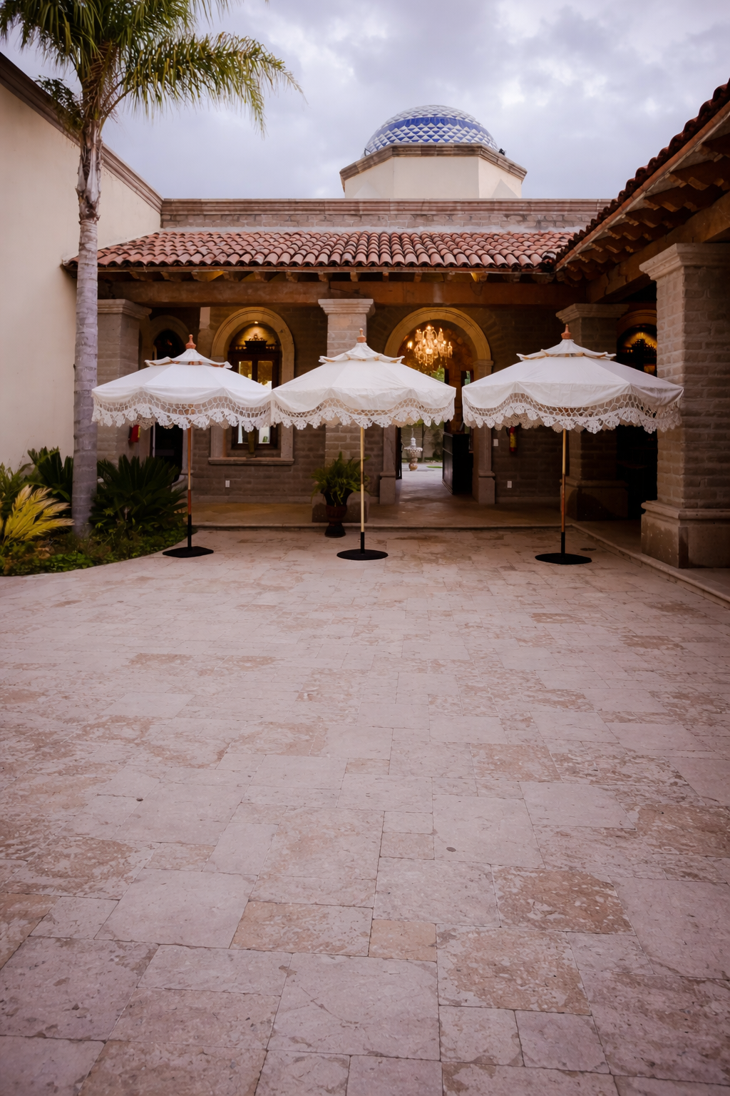 Tres sombrillas blancas con flecos se alzan sobre un patio de piedra frente a un edificio con una cúpula de azulejos azules y una palmera.