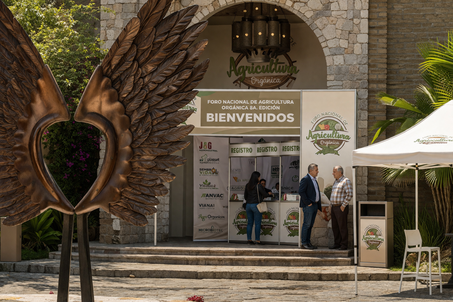 Entrada al foro agrícola Agricultura Tropical con grandes alas de bronce, señalización y gente conversando.
