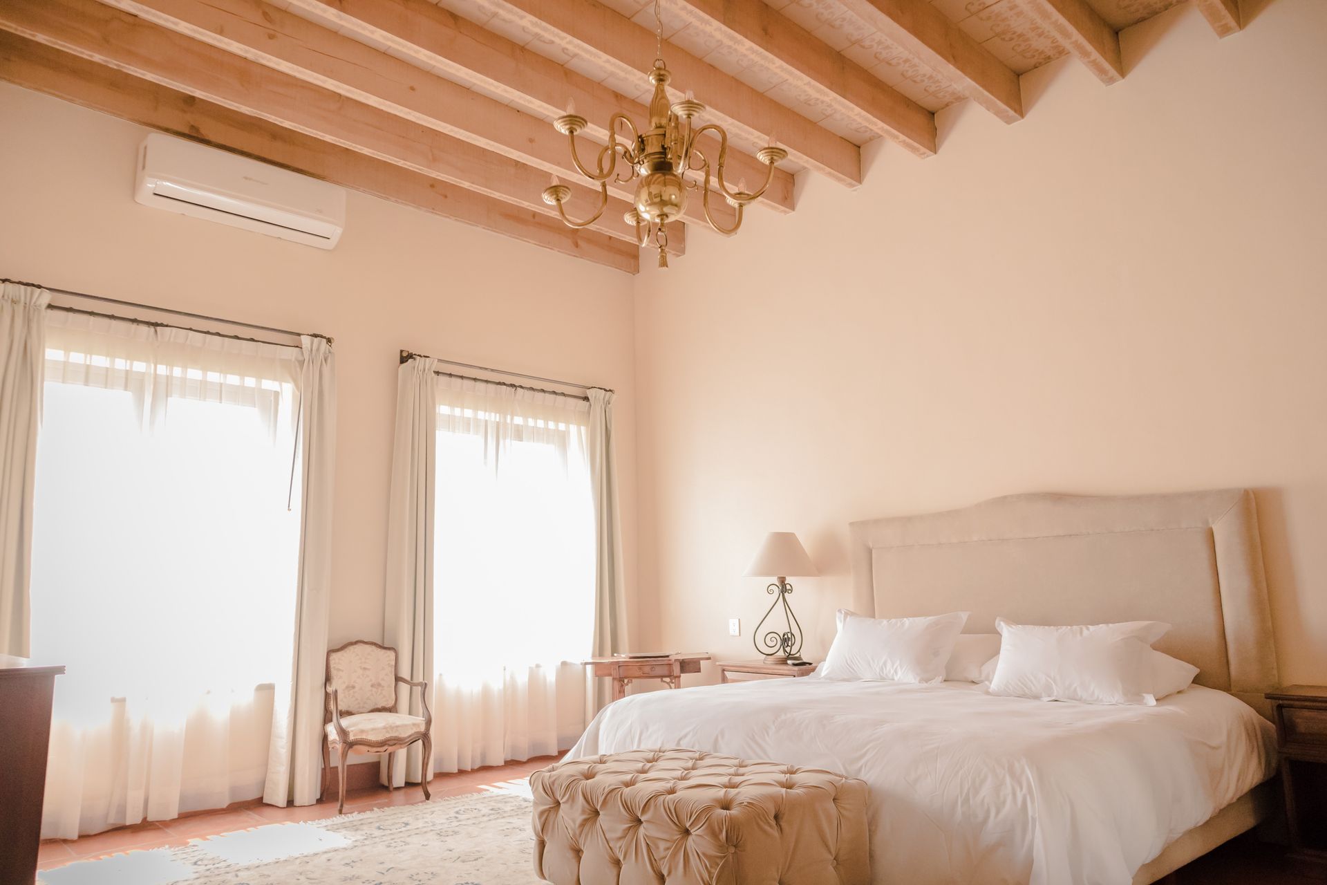 Un dormitorio luminoso de tonos neutros con vigas de madera a la vista, una lámpara de araña, dos ventanas con cortinas blancas y una cama grande.