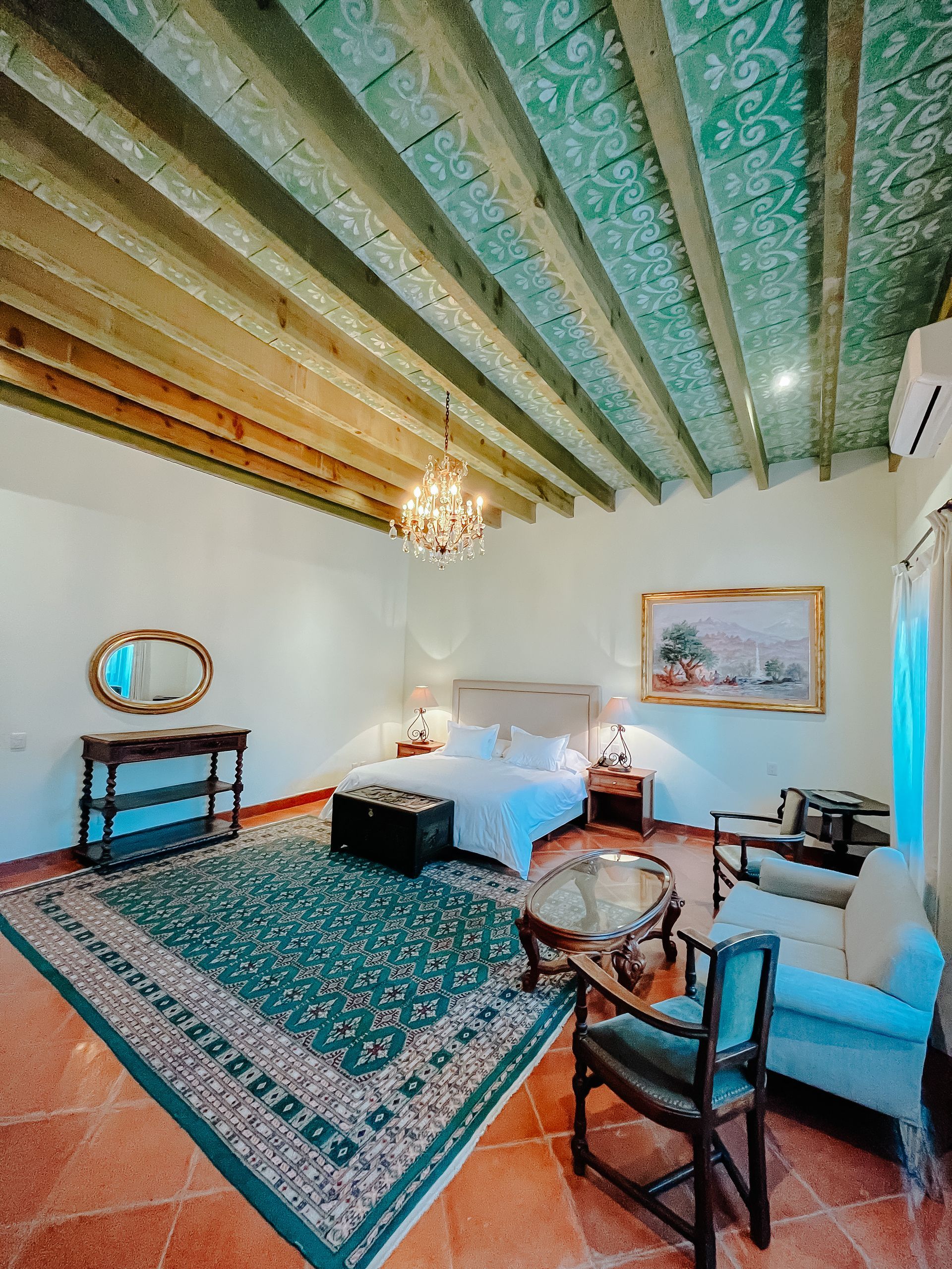 Un dormitorio con techo verde estampado, paredes blancas, una cama king size, una gran alfombra verde y sillas a juego sobre un suelo de baldosas.