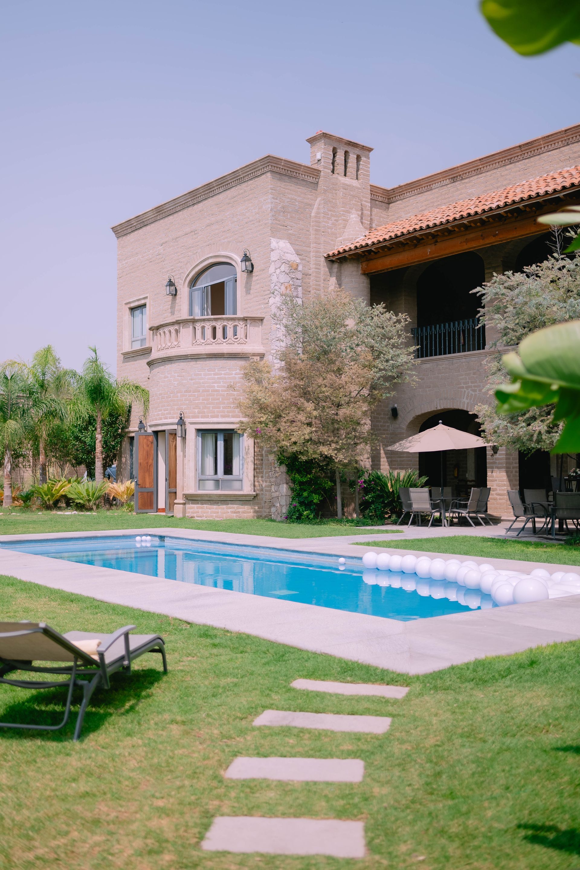 Una piscina con flotadores blancos frente a una villa de piedra de varios pisos, de estilo mediterráneo, en un día soleado.
