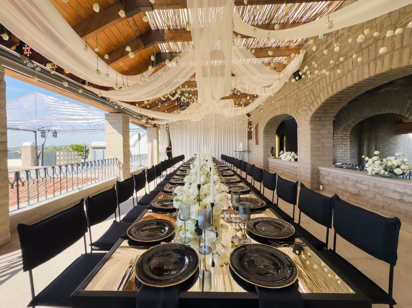 Una larga mesa preparada para un evento formal en un espacio exterior con paredes de piedra, decorada con cortinas blancas y vajilla negra.