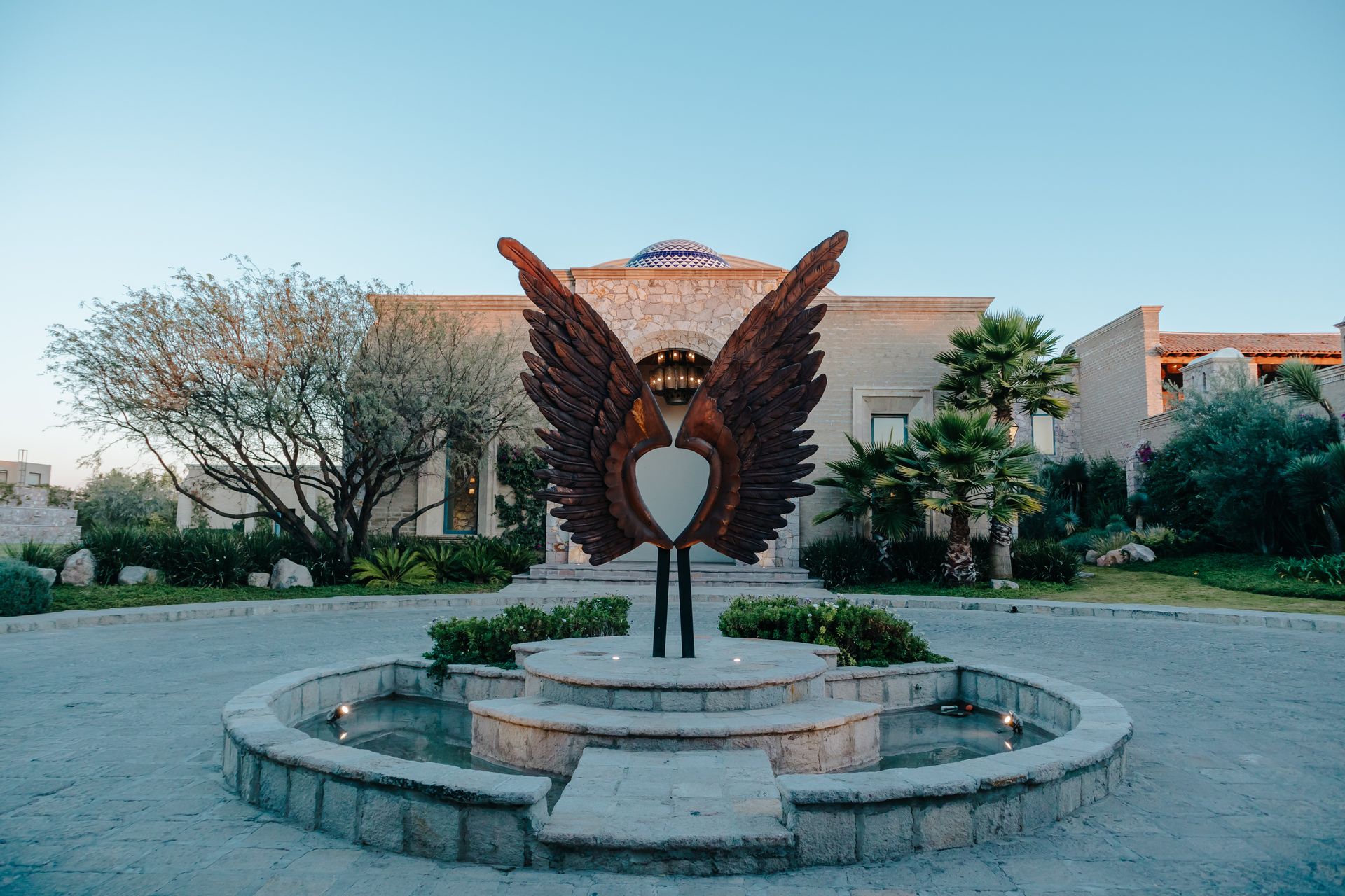 Una gran escultura de metal oxidado con forma de alas de ángel se alza en el centro de una fuente de piedra frente a una villa rústica.