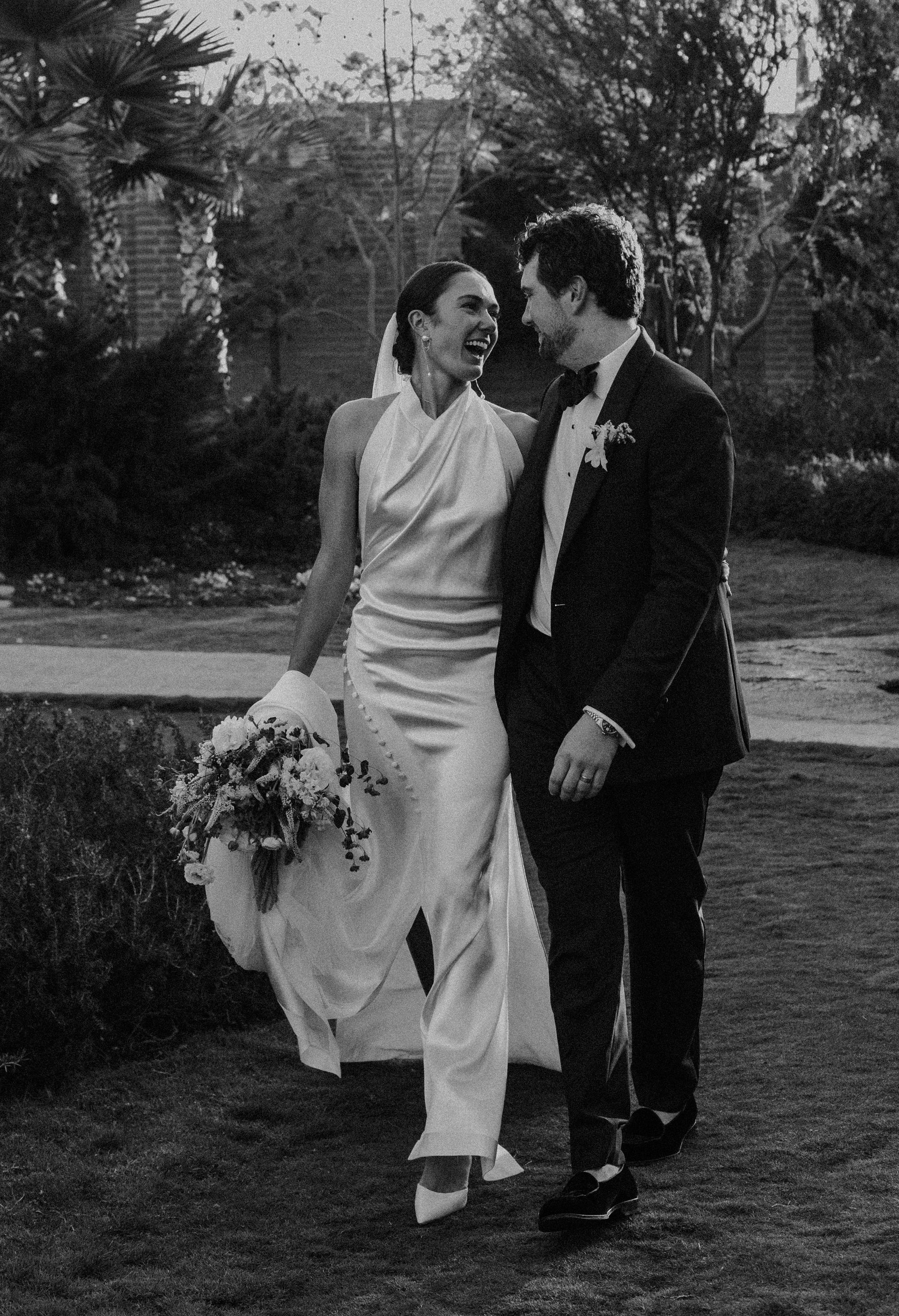 Una pareja pasea y ríe al aire libre; la persona vestida de blanco sostiene un ramo de flores.