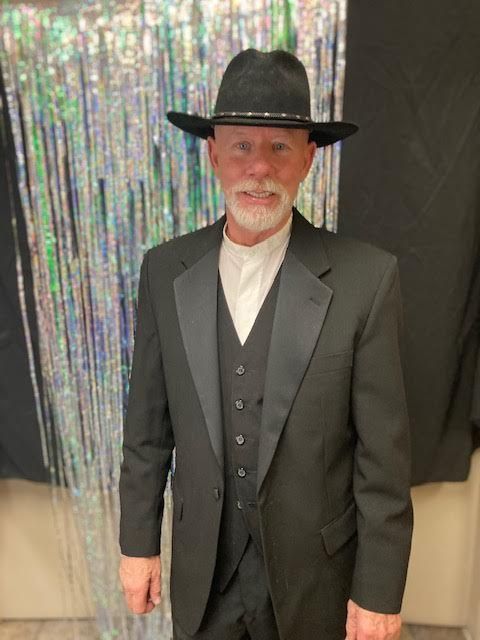 A man in a black shirt and cowboy hat is standing under a wooden arch.