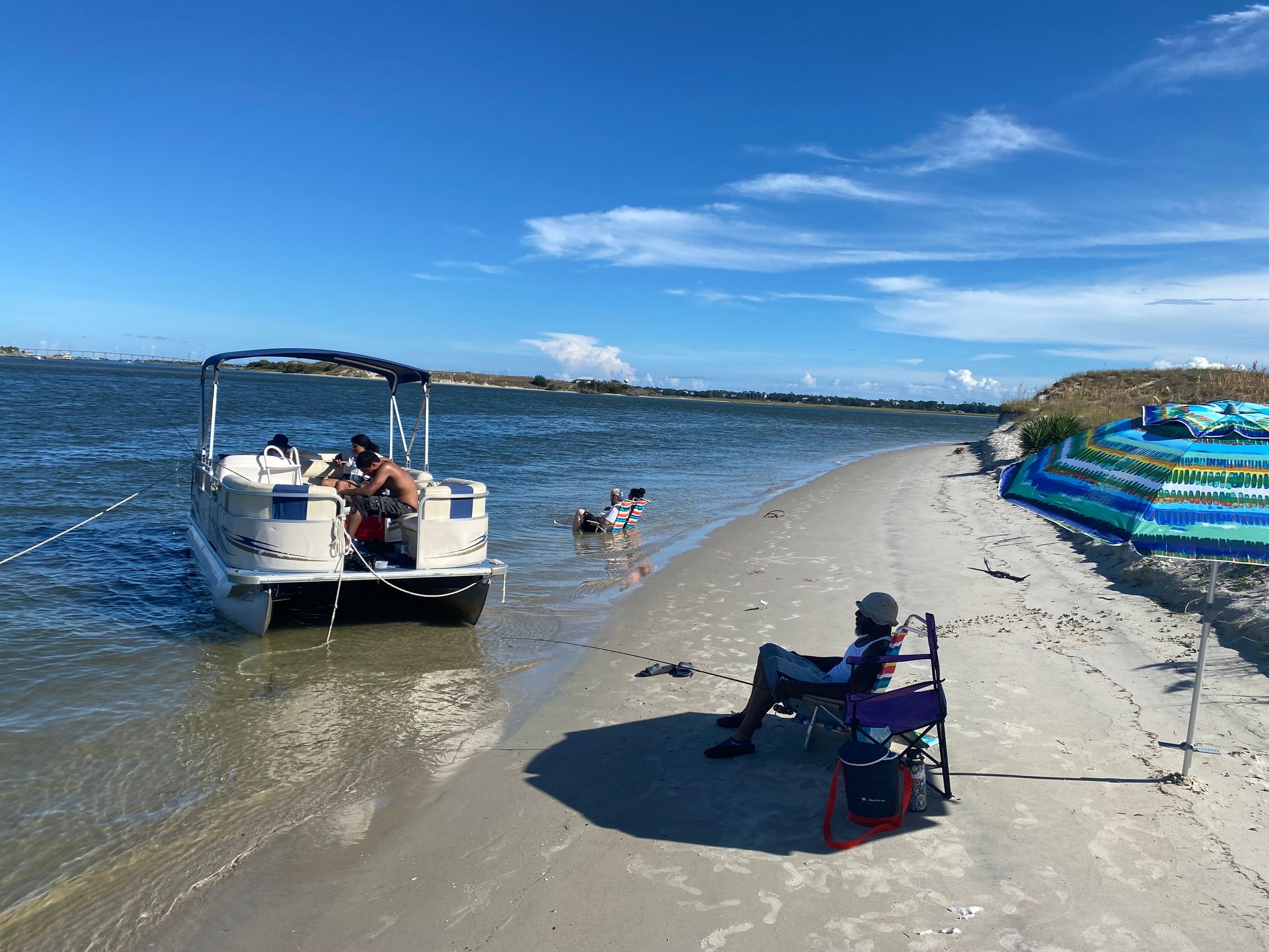 Boat near a sandy beach with people swimming, a folding chair on shore, and striped umbrellas under a blue sky