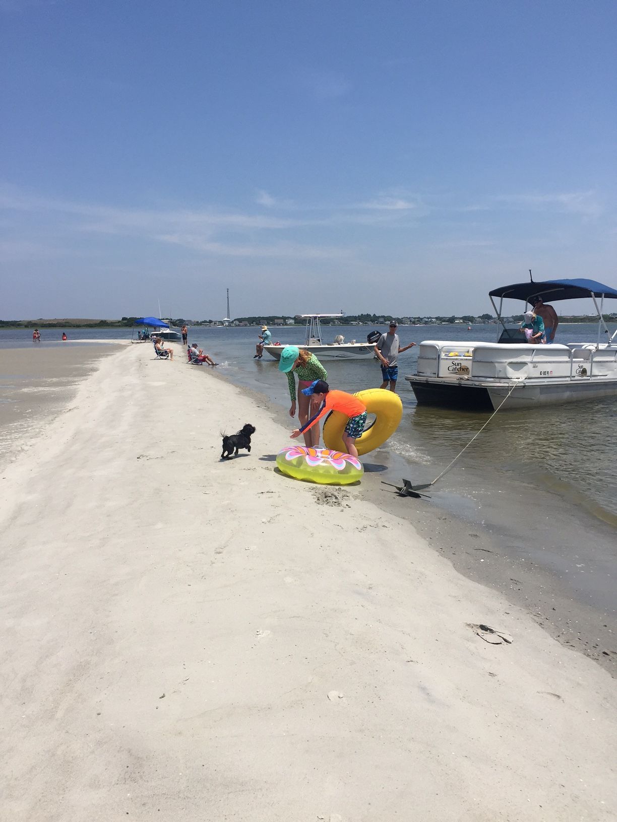 Boat near a sandy beach with people swimming, a folding chair on shore, and striped umbrellas under a blue sky