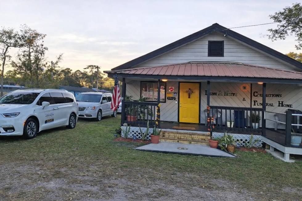 Small white house with a yellow door and porch, parked white SUVs, and an American flag at dusk
