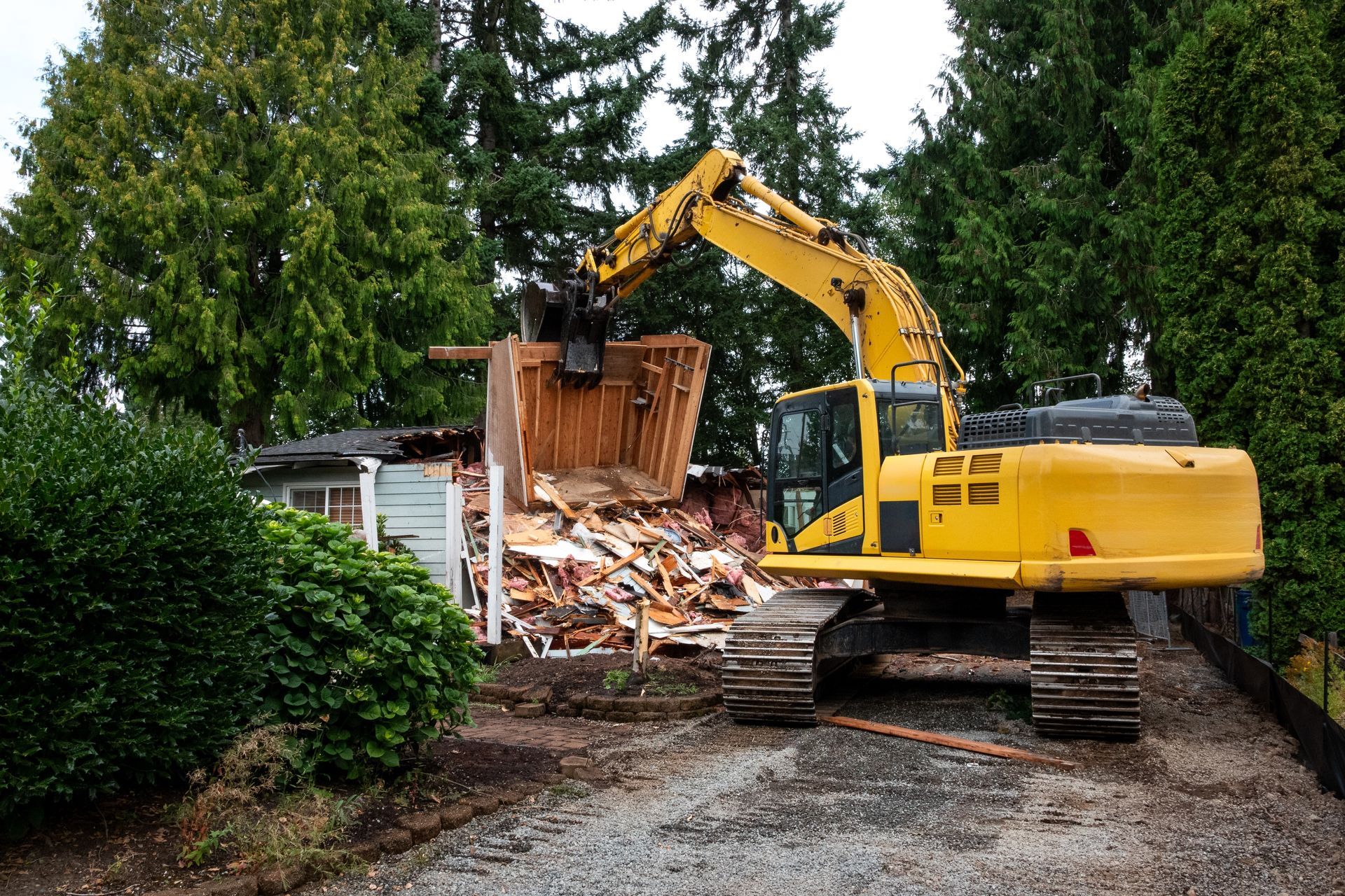 Yellow excavator demolishing a wooden structure, with debris piled beside a house and trees in the background