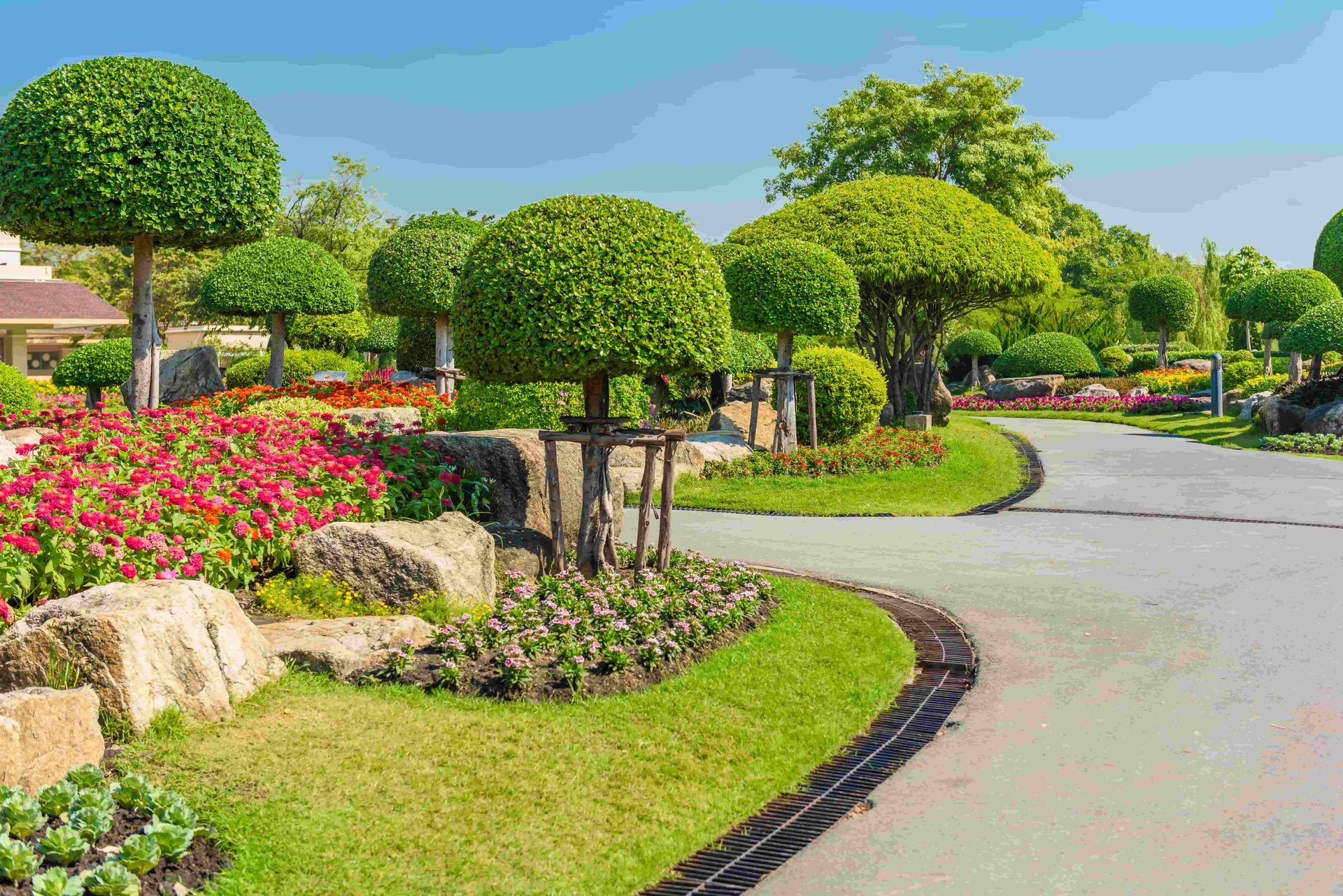 Curved park path with trimmed trees, bright flower beds, and green lawns under a clear blue sky