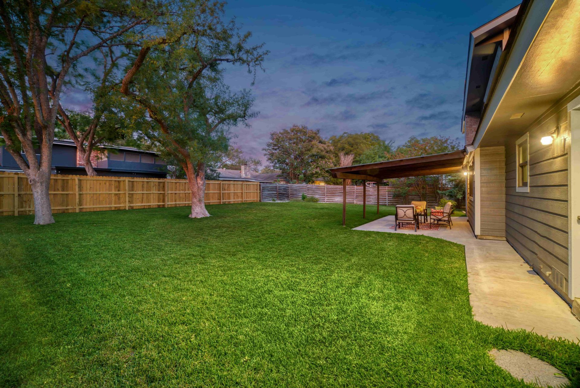 Backyard lawn at dusk beside a house, with a lit patio and trees