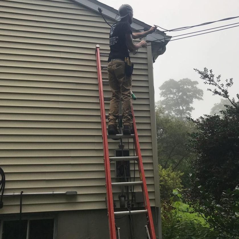 Person on a ladder, working on wires near a house. Gray siding, overcast day.