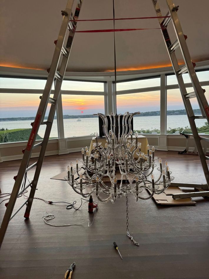 Two ladders supporting chandelier installation in a room with a waterfront view at sunset.