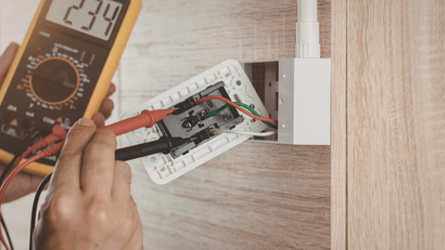 Person using a multimeter to test exposed electrical wiring in a wall outlet.
