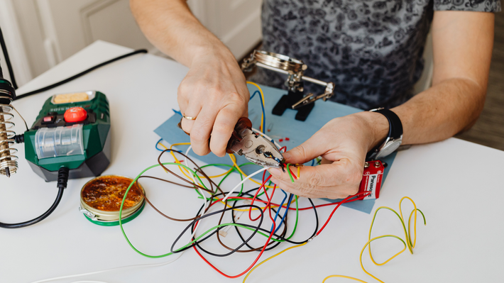 Person using pliers to work with tangled wires on a white table. Soldering iron and magnifying glass visible.
