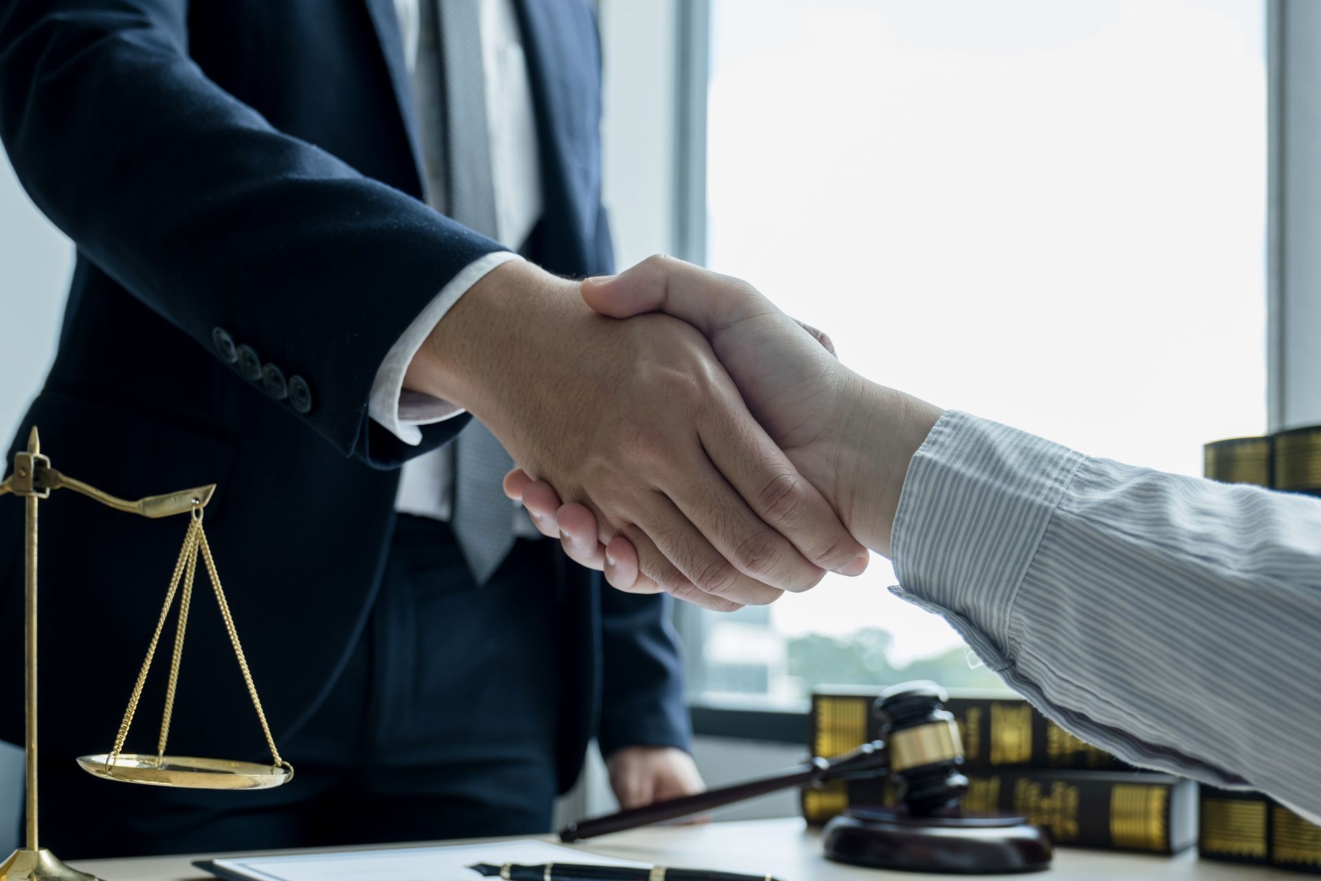 Two people shaking hands near a scale, gavel, and law books in an office setting.