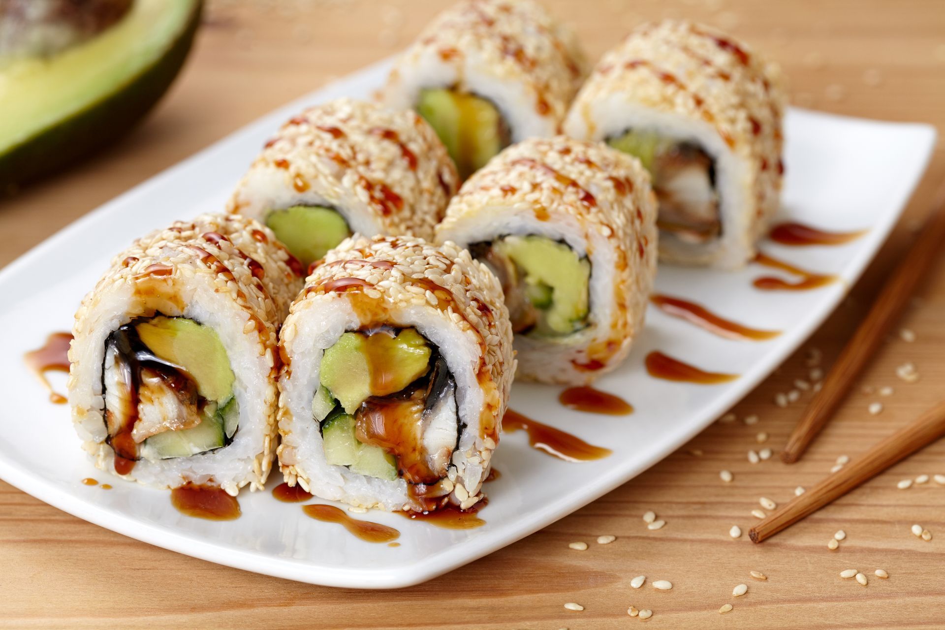 Sushi rolls topped with sesame seeds on a white plate, drizzled with sauce, with chopsticks nearby