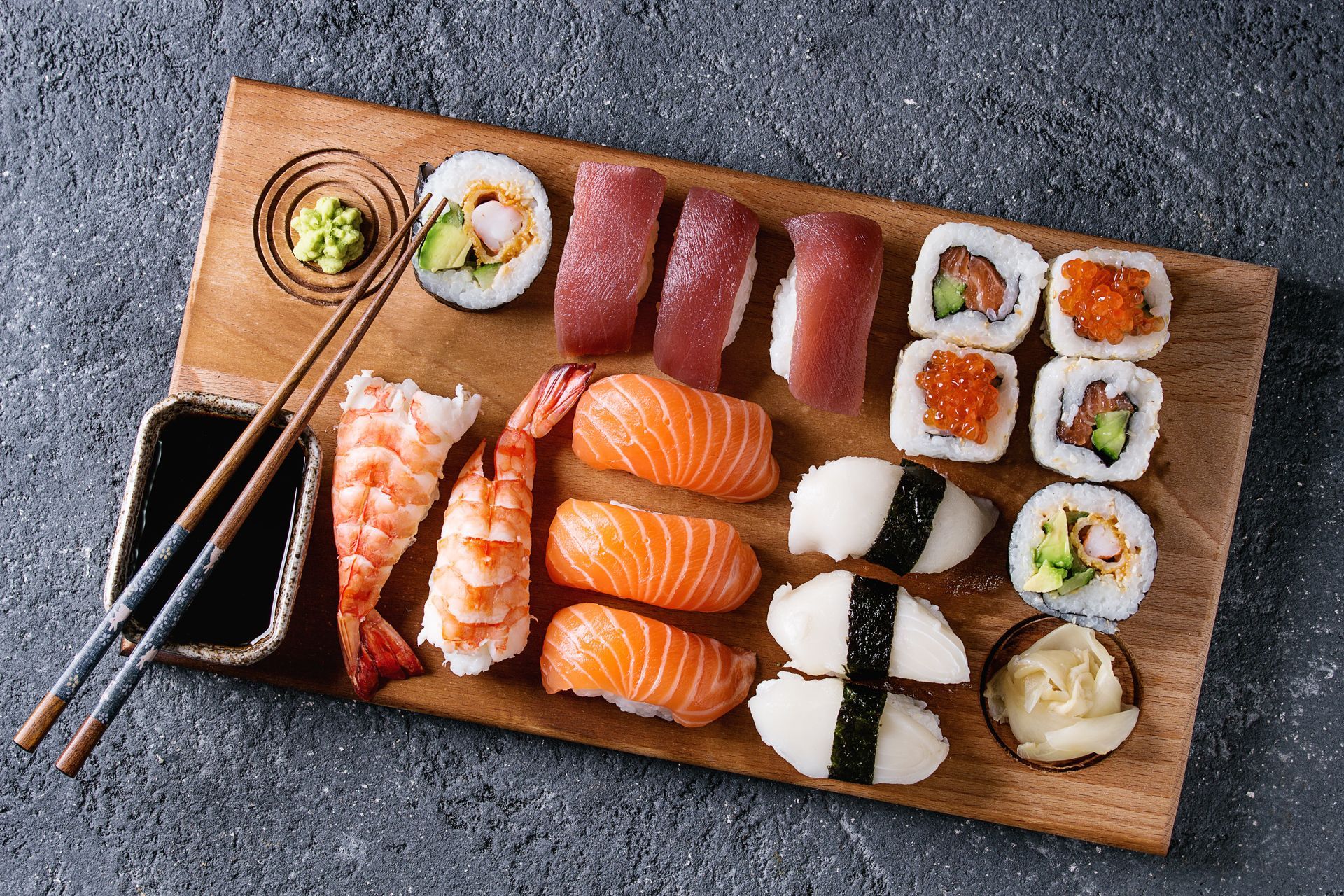 Assorted sushi rolls and nigiri on a wooden board with chopsticks and dipping sauce