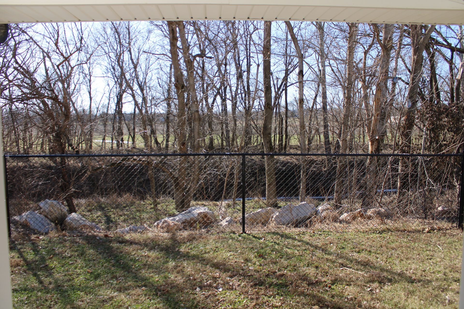 View of a black chain-link fence separating a grassy yard from a wooded area with bare trees.