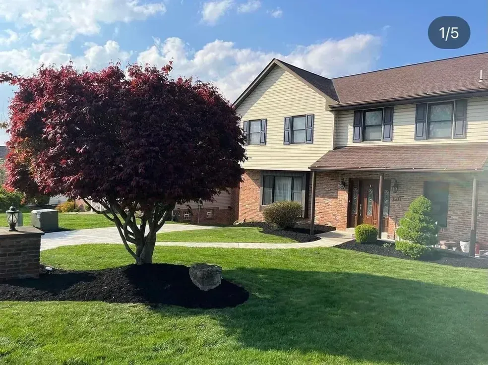 Two-story house with tan siding, brick base, and red tree in front yard on a sunny day.