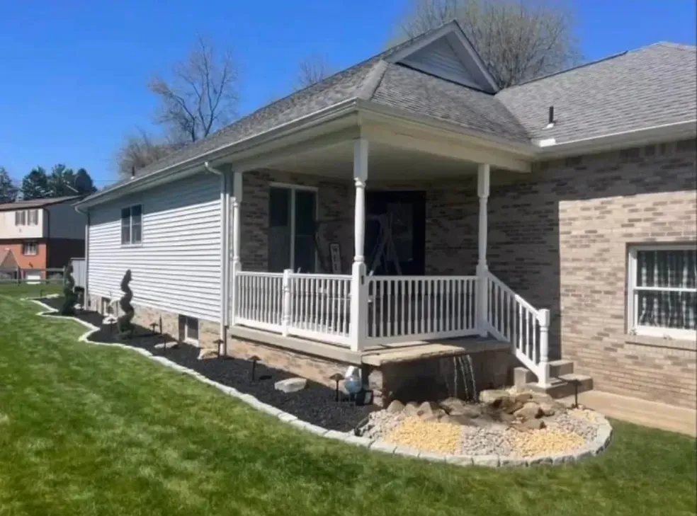 House exterior with porch, white railing, brick, and light blue siding, waterfall feature in the yard.