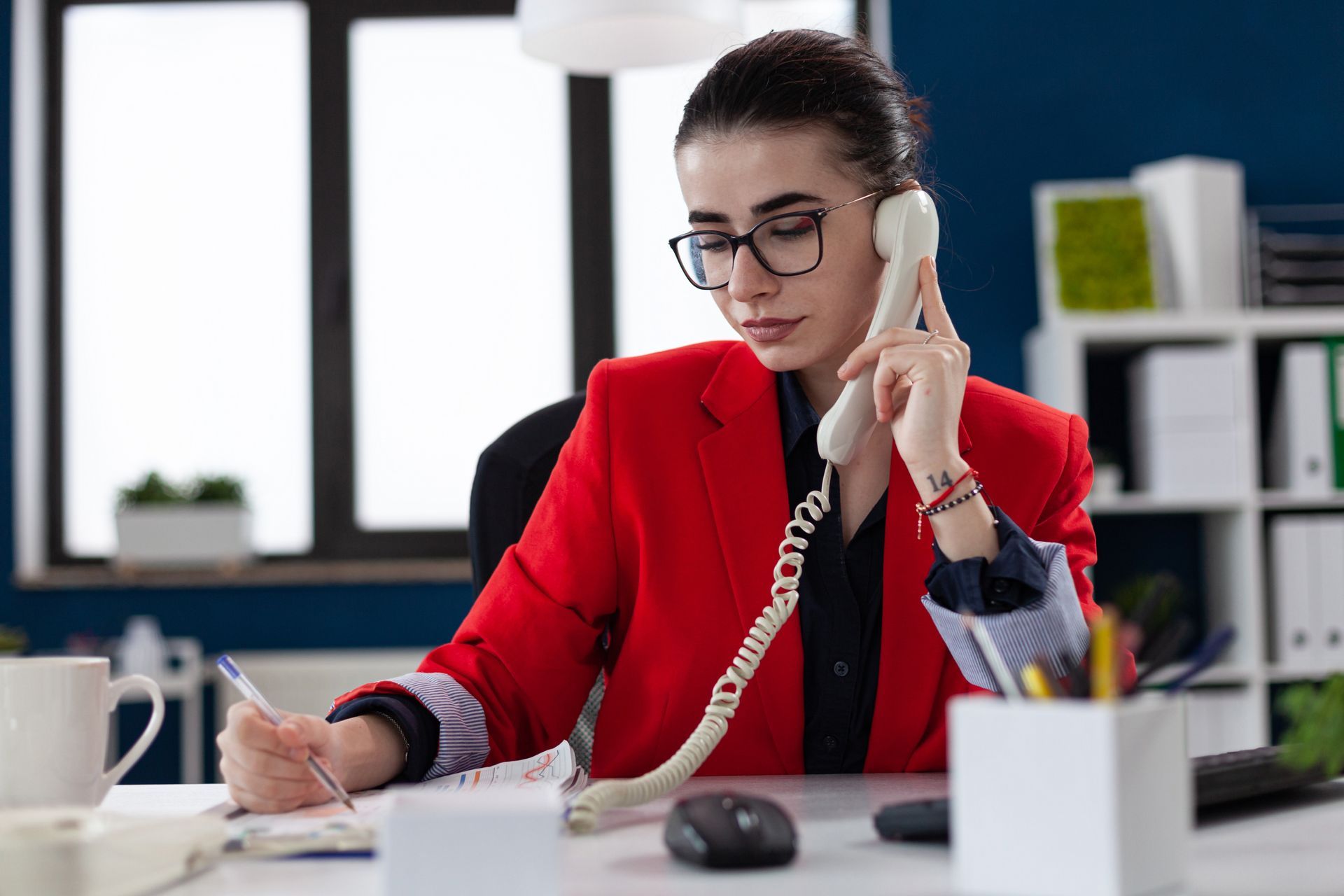 Woman in red blazer on landline phone, writing at a desk in an office. Support services in Halfmoon