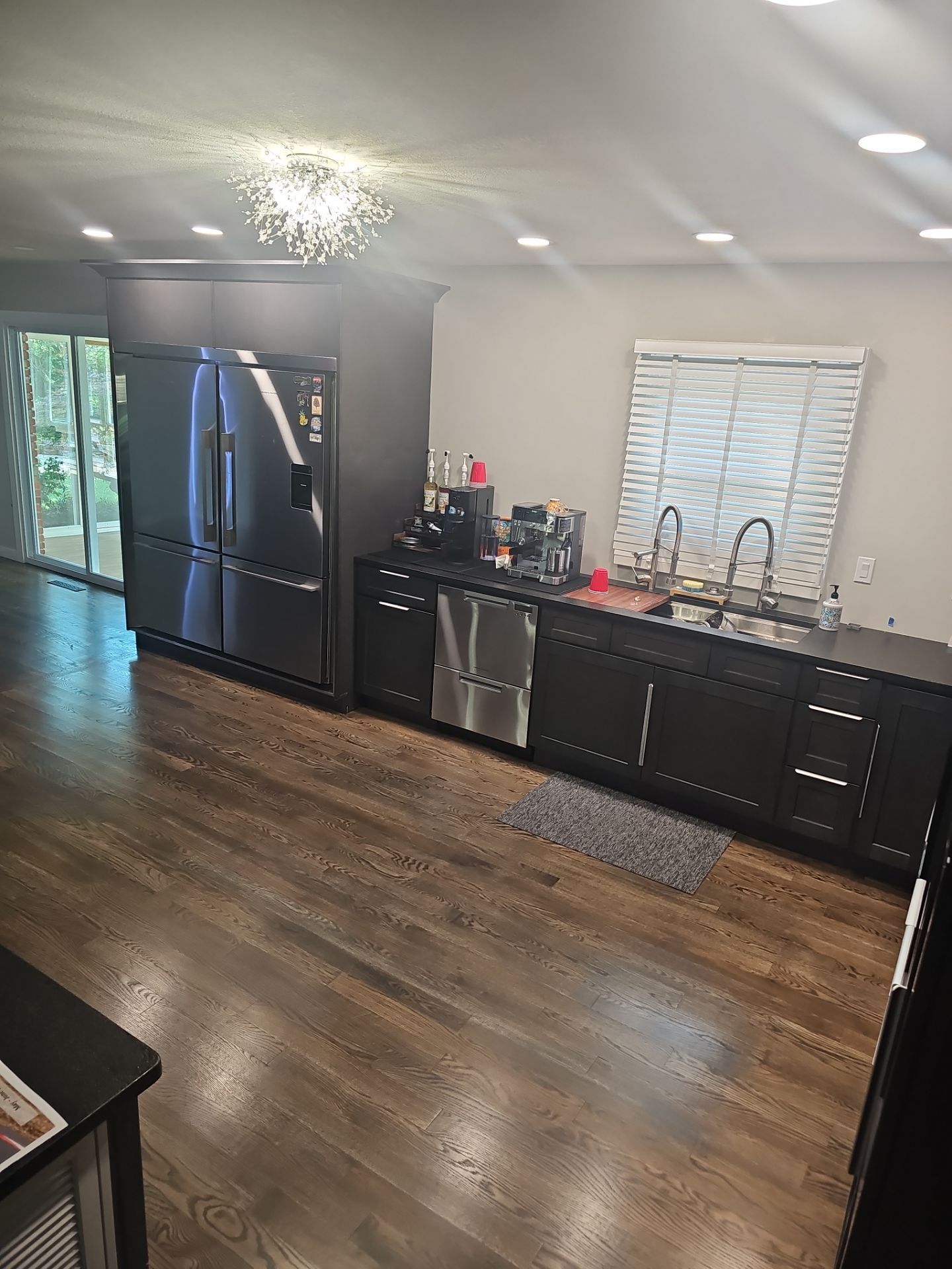 Bright kitchen with black appliances, dark cabinets, wood floors, and a sink beneath a window.