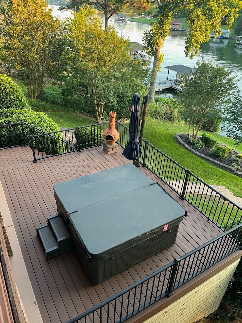 Covered hot tub on a wooden deck with black railing, overlooking a green yard and water at sunset.