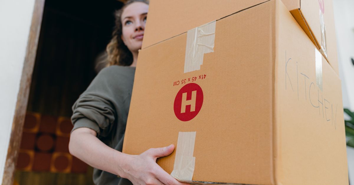 Persona que lleva una caja de cartón grande marcada con una “H” roja en una puerta.